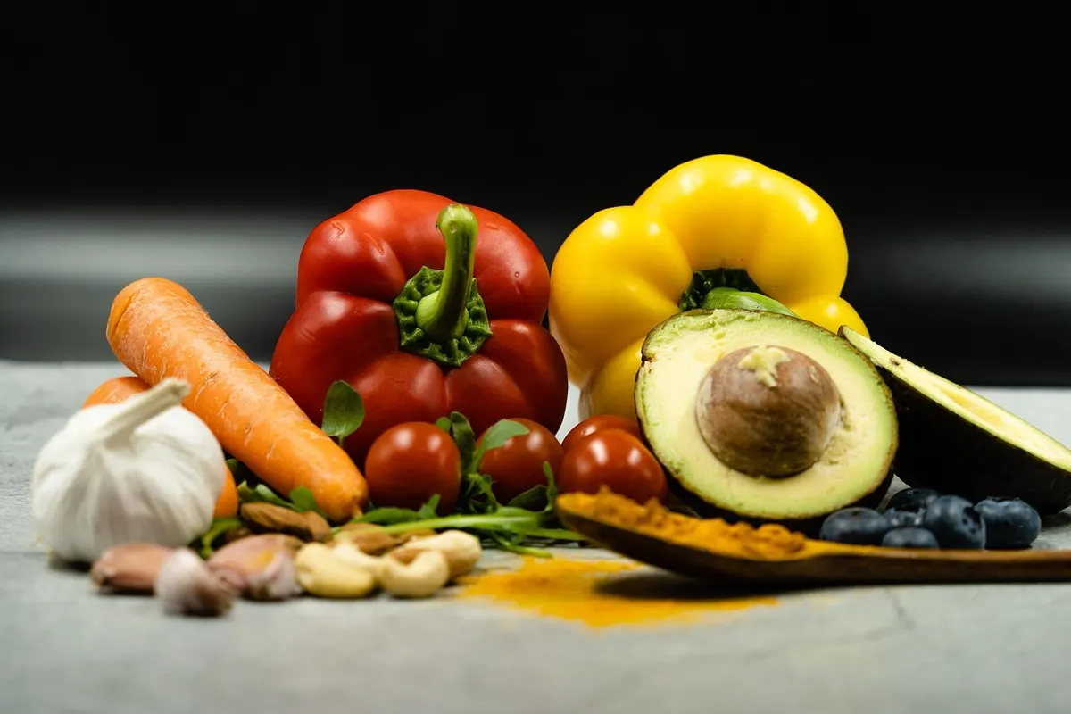 Red and yellow peppers, carrot, garlic, tomatoes, cashews, avocado halves and blueberries arranged on a grey surface