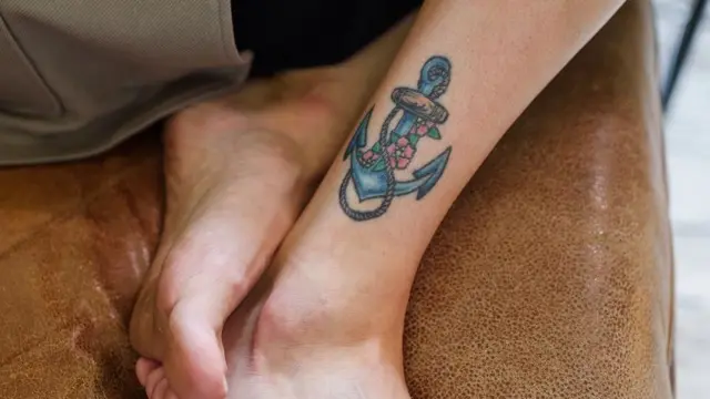 Close-up of a coloured anchor tattoo on the ankle of a bare leg, with feet crossed on a leather seat