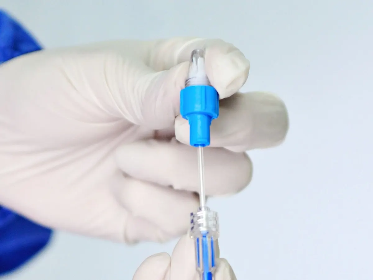 Gloved hand holding a thin medical catheter with a blue connector hub against a pale background