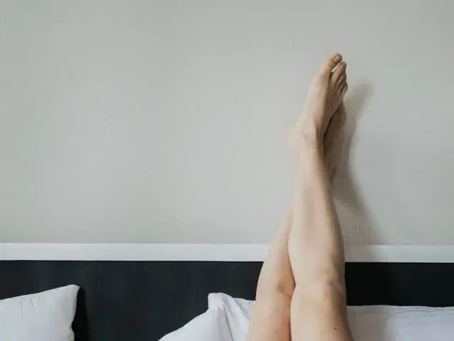 Bare legs raised and crossed at the ankles against a plain wall above a bed with white pillows
